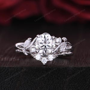 May include: A silver engagement ring with a large, round, clear center stone. The ring features a floral design with smaller accent stones and leaf-like details. The ring is set against a dark background.