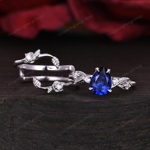 May include: A set of three silver rings. One ring features a pear-shaped, deep blue gemstone. The other two rings have a floral and leaf design with small, clear gemstones. The rings are displayed on a dark wooden surface.