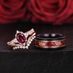 May include: A pair of wedding bands. The woman's ring has a rose gold band with a large oval ruby gemstone surrounded by smaller diamonds. The man's ring is a black band with a red and gold inlay.