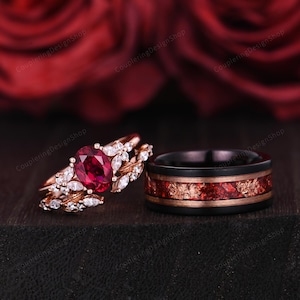 Könnte beinhalten: Ein Set aus zwei Eheringen. Der Verlobungsring hat einen großen ovalen Rubin, der von kleineren Diamanten umgeben ist, die in eine Roségold-Band gefasst sind. Der Ehering ist aus schwarzem Wolframcarbid mit roten und goldenen Akzenten.