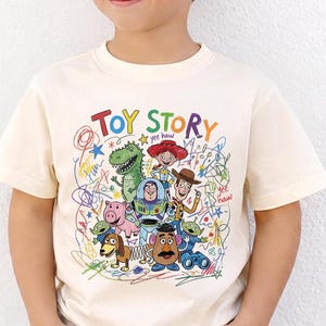 May include: Cream-colored t-shirt featuring the words "TOY STORY" in colorful letters, along with cartoon characters and scribbled designs. The shirt is worn by a young person wearing black sunglasses.