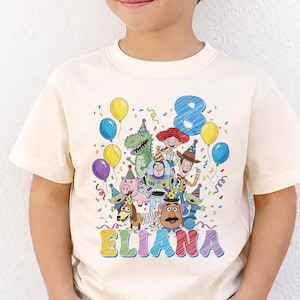 May include: Cream-colored t-shirt featuring a colorful graphic of Toy Story characters, balloons, and confetti. The number "8" and the name "ELIANA" are also displayed. The shirt is worn by a child wearing black sunglasses.