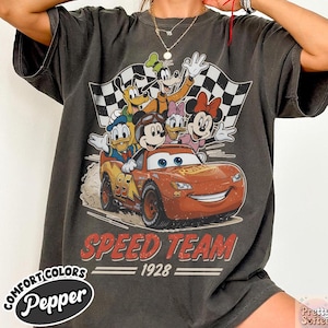 K&ouml;nnte beinhalten: Dunkelgraues T-Shirt mit einer Grafik von Disney-Figuren, darunter Micky Maus und Donald Duck, mit einem roten Rennwagen. Der Text "SPEED TEAM 1928" ist unter dem Bild aufgedruckt. Das Shirt hat die Worte "Comfort Colors Pepper" aufgedruckt.