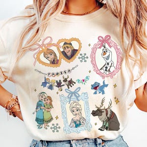 May include: Cream-colored t-shirt featuring various characters from the animated film "Frozen." The design includes framed portraits of Anna, Elsa, Kristoff, and Olaf, along with other characters and the word "FROZEN."