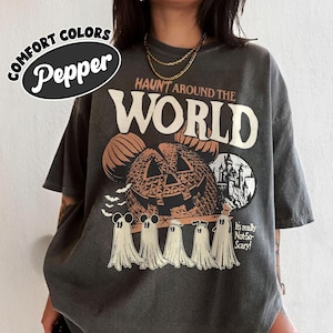 May include: Dark grey t-shirt with a Halloween-themed graphic. The shirt features the text "HAUNT AROUND THE WORLD" above a jack-o'-lantern design and cartoon ghosts. The shirt also has the text "It's really Not-So-Scary!" and the words "COMFORT COLORS Pepper".