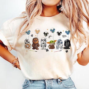 Könnte beinhalten: Cremefarbenes T-Shirt mit Cartoon-Star-Wars-Charakteren, darunter BB-8, Chewbacca, ein Sturmtruppler, Grogu, R2-D2 und Darth Vader. Jeder Charakter hält einen Ballon. Das Shirt hat einen Rundhalsausschnitt und kurze Ärmel.