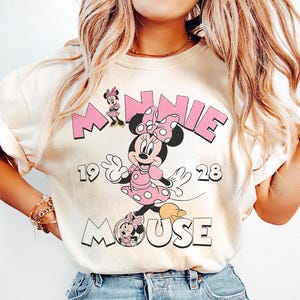 Puede incluir: Camiseta color crema con un gr&aacute;fico de Minnie Mouse con un vestido de lunares rosa y un lazo. El texto "MINNIE 1928 MOUSE" est&aacute; impreso en rosa y blanco. El dise&ntilde;o es de estilo vintage y de dibujos animados.