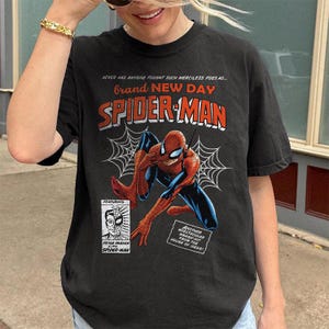 K&ouml;nnte beinhalten: Schwarzes T-Shirt mit einer Spider-Man-Comic-Grafik. Das Design enth&auml;lt die W&ouml;rter "Brand New Day" und "Spider-Man" in Rot und Wei&szlig;, zusammen mit Spinnweben und zus&auml;tzlichem Text.