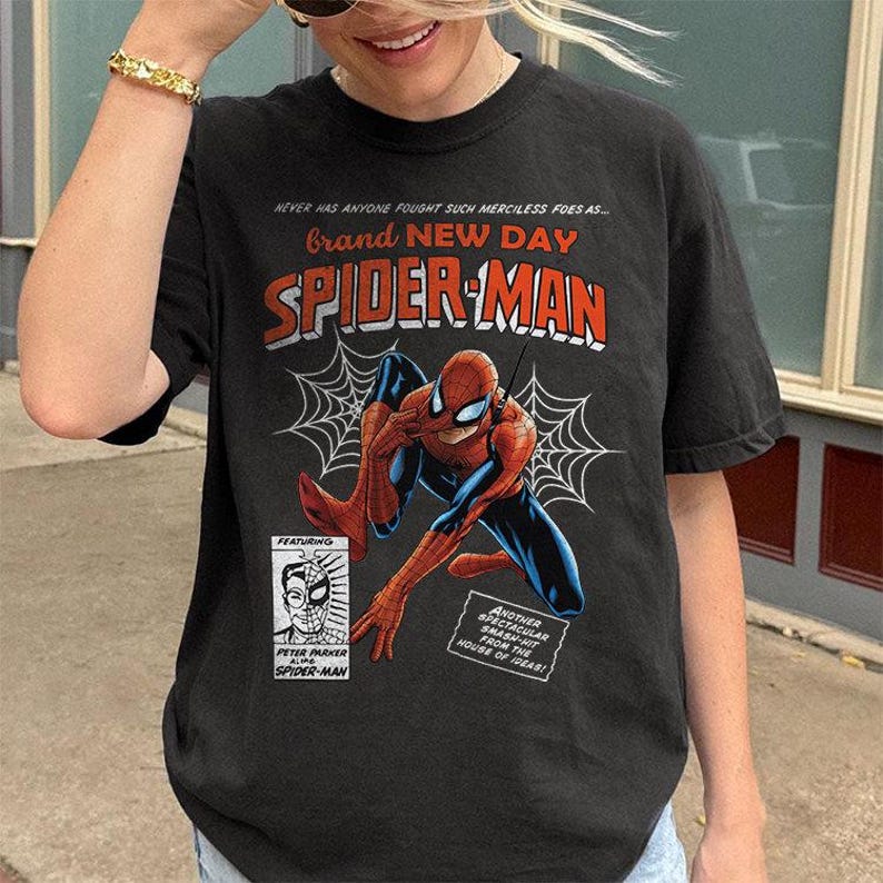 K&ouml;nnte beinhalten: Schwarzes T-Shirt mit einer Spider-Man-Comic-Grafik. Das Design enth&auml;lt die W&ouml;rter "Brand New Day" und "Spider-Man" in Rot und Wei&szlig;, zusammen mit Spinnweben und zus&auml;tzlichem Text.