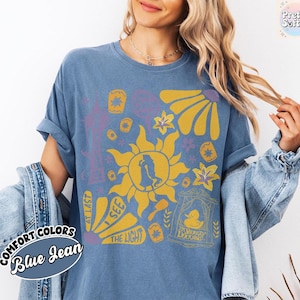 K&ouml;nnte beinhalten: Blaues Jeans-T-Shirt mit einem gelben Sonnendesign, einer Silhouette und floralen Akzenten. Auf dem Shirt stehen die Worte "At Last I See The Light" und "The Snuggly Duckling". Teil der Comfort Colors Kollektion.