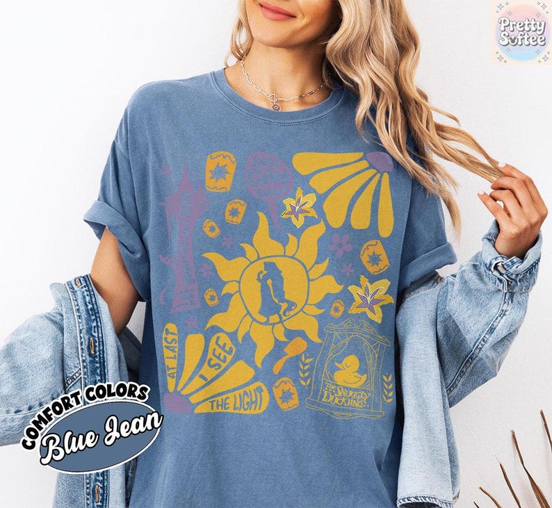 K&ouml;nnte beinhalten: Blaues Jeans-T-Shirt mit einem gelben Sonnendesign, einer Silhouette und floralen Akzenten. Auf dem Shirt stehen die Worte "At Last I See The Light" und "The Snuggly Duckling". Teil der Comfort Colors Kollektion.