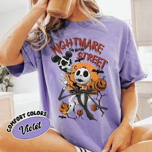 May include: Violet Comfort Colors t-shirt featuring a Halloween-themed graphic. The design includes Jack Skellington, pumpkins, bats, and the text "Nightmare on Main Street." A Mickey Mouse skull is also present in the design.