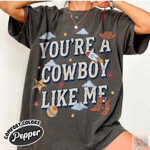May include: A charcoal gray short-sleeve t-shirt featuring the text "YOU'RE A COWBOY LIKE ME" in large white letters with blue outlines. The design includes illustrations of a brown cowboy hat, a rocket, a sheriff star, boots, and clouds.