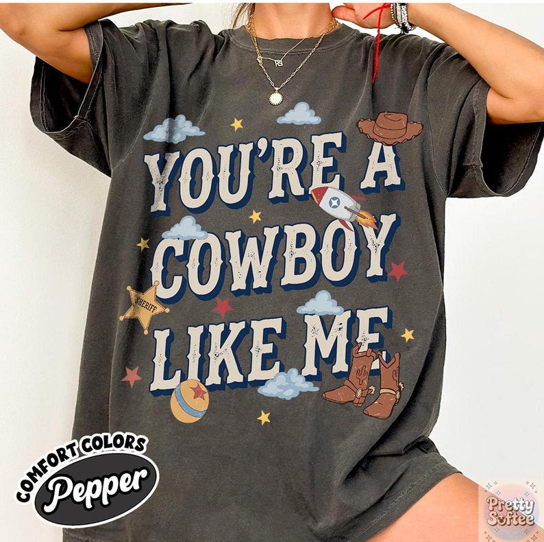 May include: A charcoal gray short-sleeve t-shirt featuring the text "YOU'RE A COWBOY LIKE ME" in large white letters with blue outlines. The design includes illustrations of a brown cowboy hat, a rocket, a sheriff star, boots, and clouds.