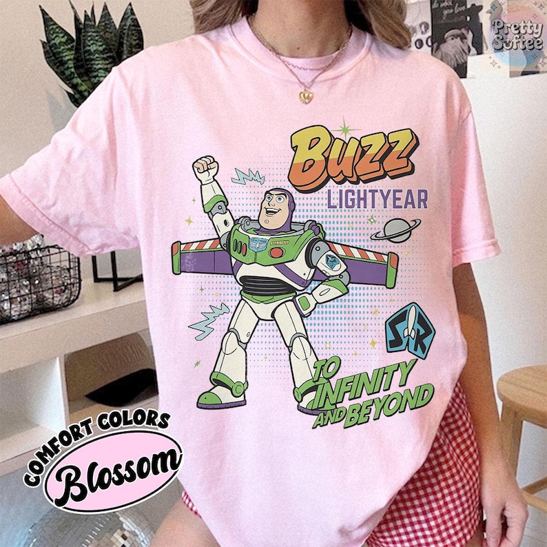 May include: A pale pink t-shirt with a graphic of Buzz Lightyear, the words "Buzz Lightyear" and "To Infinity and Beyond." The shirt also has the text "Comfort Colors Blossom."