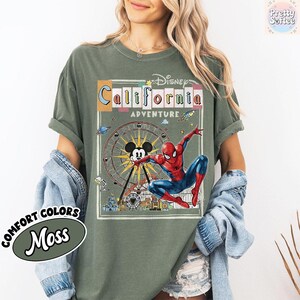 Puede incluir: Camiseta verde musgo con un gr&aacute;fico de una noria, Mickey Mouse y Spider-Man. El texto "Disney California Adventure" se muestra encima del gr&aacute;fico. La camiseta tambi&eacute;n tiene las palabras "Comfort Colors" y "Moss" en la parte inferior izquierda.