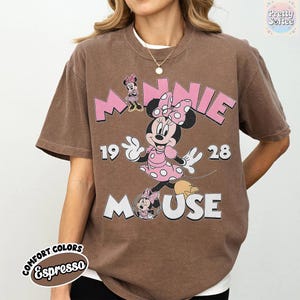 Puede incluir: Camiseta color espresso con un dise&ntilde;o de Minnie Mouse. El dise&ntilde;o incluye las palabras "MINNIE MOUSE" en rosa, junto con los n&uacute;meros "1928". La camiseta tiene un aspecto vintage, ligeramente descolorido.