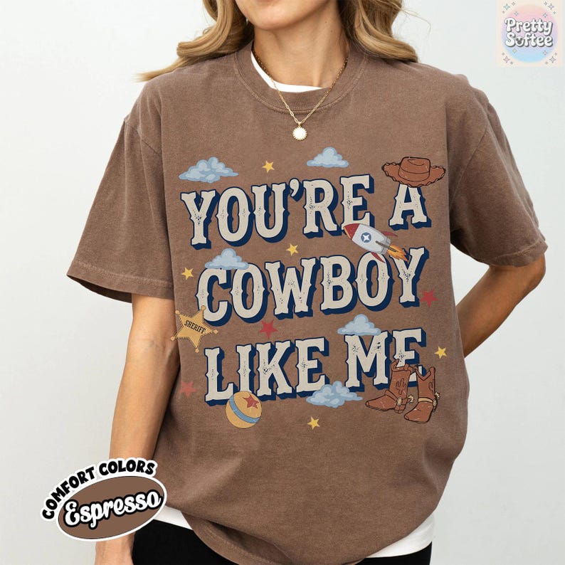 May include: An espresso brown short-sleeve t-shirt featuring the text "YOU'RE A COWBOY LIKE ME" in a bold, dark blue font. The graphic includes illustrations of a cowboy hat, a rocket, a sheriff star, a ball, and cowboy boots with clouds and stars.