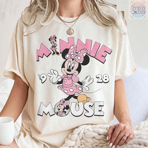 Puede incluir: Camiseta color crema con un gr&aacute;fico de Minnie Mouse con un vestido y lazo de lunares rosas. La palabra "MINNIE" est&aacute; en letras rosas en bloque sobre el personaje, con "MOUSE" debajo. Tambi&eacute;n son visibles los n&uacute;meros "1928".
