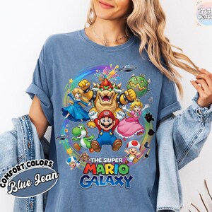 K&ouml;nnte beinhalten: Blaues Jeans-T-Shirt mit farbenfroher Grafik von Super Mario Galaxy-Charakteren. Das Design zeigt Mario, Prinzessin Peach, Bowser und Yoshi, mit dem Text "THE SUPER MARIO GALAXY".