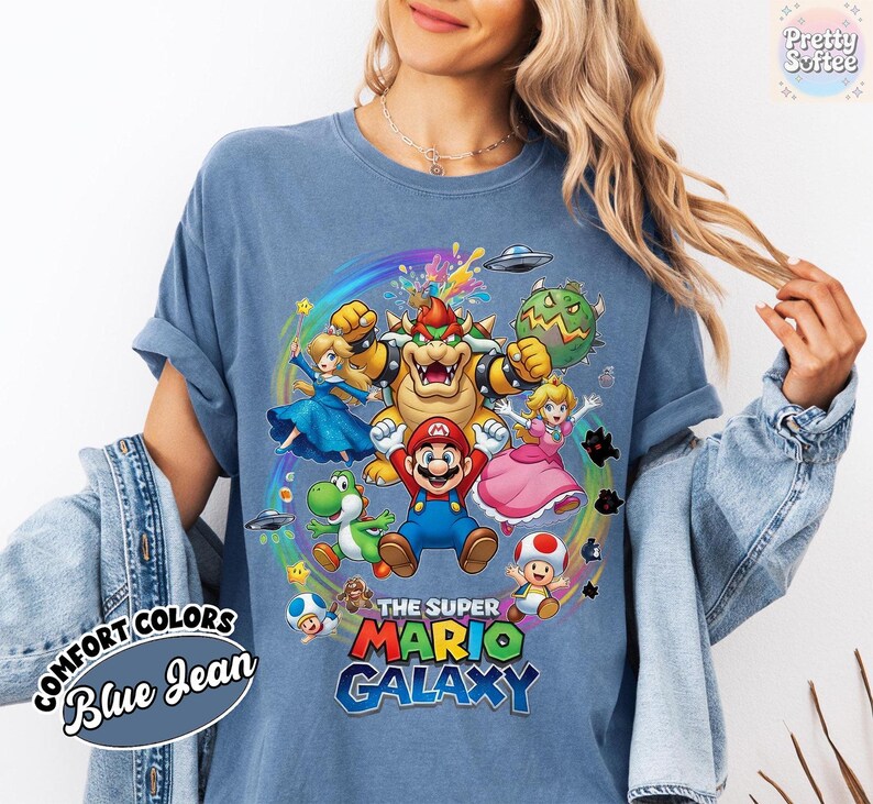 K&ouml;nnte beinhalten: Blaues Jeans-T-Shirt mit farbenfroher Grafik von Super Mario Galaxy-Charakteren. Das Design zeigt Mario, Prinzessin Peach, Bowser und Yoshi, mit dem Text "THE SUPER MARIO GALAXY".