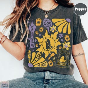 K&ouml;nnte beinhalten: Dunkelgraues T-Shirt mit einem farbenfrohen Grafikdesign mit einer Sonne, Blumen und Text, der "Best Day Ever", "I See The Light" und "Let Your Power Shine" lautet. Das Shirt hat auch eine Silhouette eines Paares.