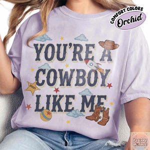 May include: A light purple t-shirt featuring the dark blue text YOU'RE A COWBOY LIKE ME. The design includes a brown cowboy hat, a rocket ship, a yellow sheriff star, a colorful ball, brown cowboy boots, small red stars, and light blue clouds.