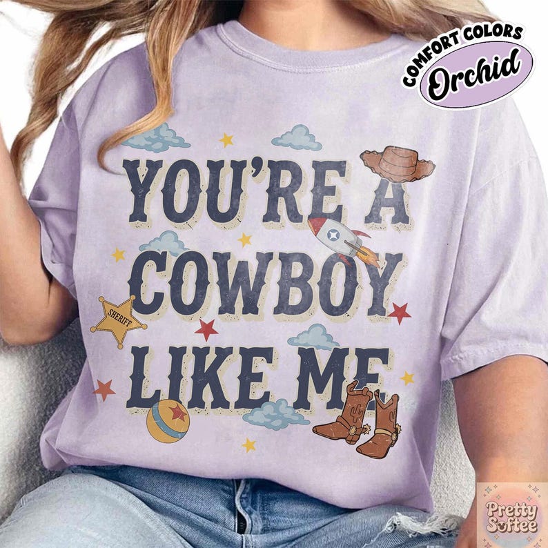 May include: A light purple t-shirt featuring the dark blue text YOU'RE A COWBOY LIKE ME. The design includes a brown cowboy hat, a rocket ship, a yellow sheriff star, a colorful ball, brown cowboy boots, small red stars, and light blue clouds.