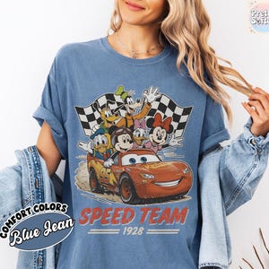 K&ouml;nnte beinhalten: Blaues Jeans-T-Shirt mit einer Grafik von Disney-Figuren in einem Rennwagen. Das Design umfasst Micky Maus, Donald Duck, Goofy sowie Minnie und Daisy Duck. Der Text "SPEED TEAM 1928" ist rot gedruckt.