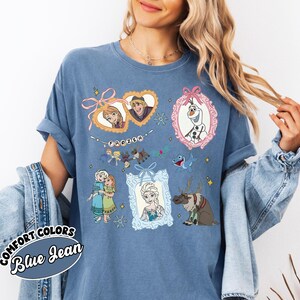 May include: Blue jean-colored t-shirt featuring illustrations of characters from the movie "Frozen." The design includes framed portraits of Anna, Elsa, Kristoff, and Olaf, along with other characters and the word "Frozen."