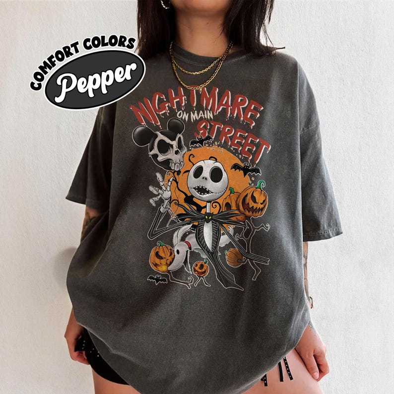 May include: Dark gray t-shirt with a graphic featuring Jack Skellington from The Nightmare Before Christmas, pumpkins, and bats. The text "Nightmare on Main Street" is printed above the character. The shirt has a distressed look and the text "Comfort Colors Pepper" is printed on the top left.