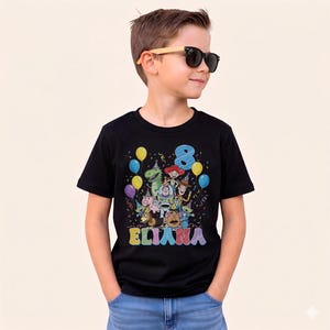 May include: A black t-shirt featuring a colorful graphic of Toy Story characters, balloons, confetti, the number 8, and the name ELIANA. The shirt is worn by a child wearing sunglasses.