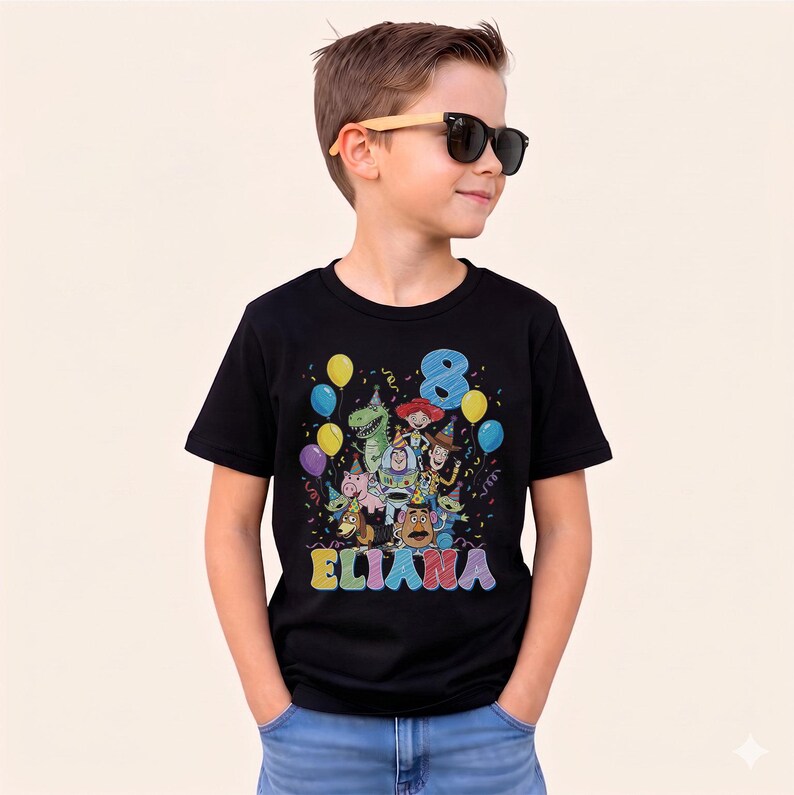 May include: A black t-shirt featuring a colorful graphic of Toy Story characters, balloons, confetti, the number 8, and the name ELIANA. The shirt is worn by a child wearing sunglasses.