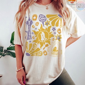 K&ouml;nnte beinhalten: Cremefarbenes T-Shirt mit gelbem Sonnendesign und einer Personensilhouette, umgeben von Grafiken und Text. Der Text enth&auml;lt "Let your Power Shine", "Best Day Ever" und "The Snuggly Duckling".