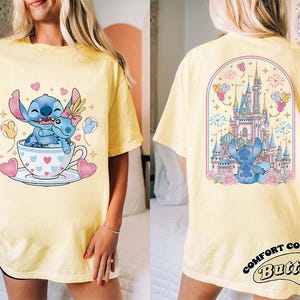 May include: Yellow t-shirt featuring a cartoon Stitch design. The front shows Stitch in a teacup with hearts and balloons. The back displays a castle scene with Stitch and fireworks. The shirt has the text "Comfort Colors Butter".