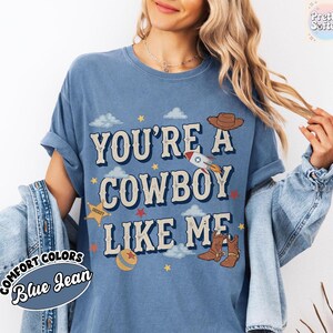 May include: A blue jean colored t-shirt featuring the text YOU'RE A COWBOY LIKE ME in large letters. The graphic includes a brown cowboy hat, a rocket ship, a yellow sheriff star, a ball, and small clouds and stars scattered across the front.