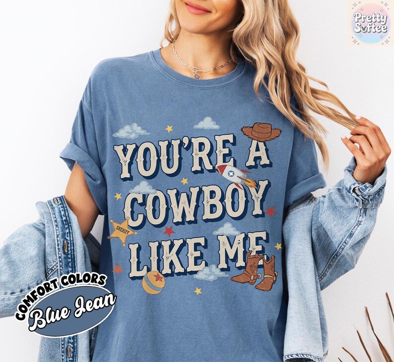 May include: A blue jean colored t-shirt featuring the text YOU'RE A COWBOY LIKE ME in large letters. The graphic includes a brown cowboy hat, a rocket ship, a yellow sheriff star, a ball, and small clouds and stars scattered across the front.