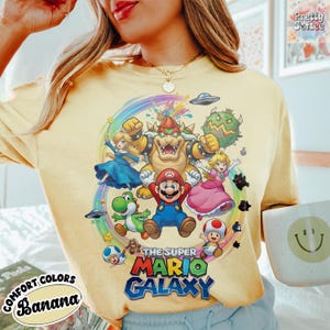 K&ouml;nnte beinhalten: Hellgelbes Comfort Colors T-Shirt mit einer farbenfrohen Grafik von Mario und anderen Charakteren aus dem Spiel Super Mario Galaxy. Der Text "THE SUPER MARIO GALAXY" ist in einer leuchtenden Schriftart gedruckt. Das Shirt hat auch den Text "COMFORT COLORS BANANA".