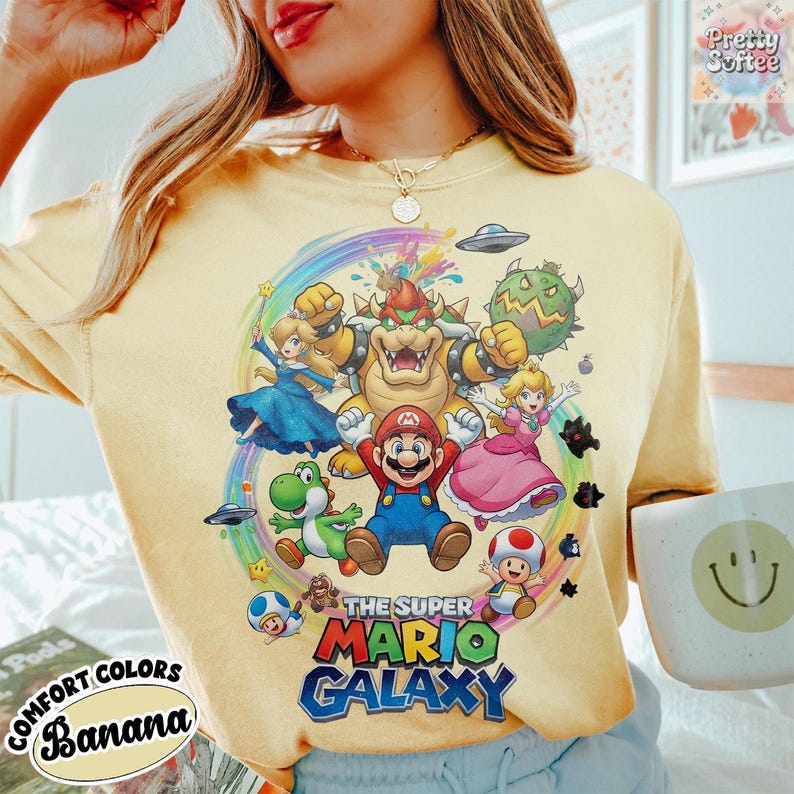 K&ouml;nnte beinhalten: Hellgelbes Comfort Colors T-Shirt mit einer farbenfrohen Grafik von Mario und anderen Charakteren aus dem Spiel Super Mario Galaxy. Der Text "THE SUPER MARIO GALAXY" ist in einer leuchtenden Schriftart gedruckt. Das Shirt hat auch den Text "COMFORT COLORS BANANA".