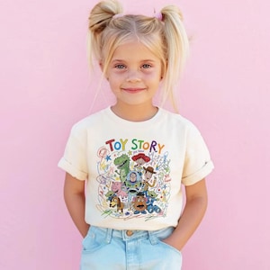 May include: A cream-colored t-shirt featuring the words "Toy Story" in colorful letters, with a graphic of characters from the movie. The shirt is worn by a young girl with blonde pigtails and blue jeans.
