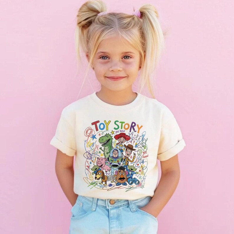 May include: A cream-colored t-shirt featuring the words "Toy Story" in colorful letters, with a graphic of characters from the movie. The shirt is worn by a young girl with blonde pigtails and blue jeans.