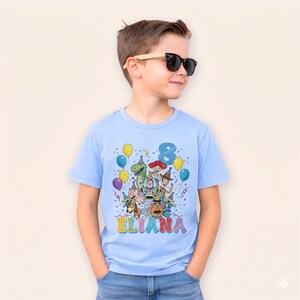May include: A light blue t-shirt featuring a colorful graphic of Toy Story characters, balloons, and confetti. The number 8 and the name "Eliana" are also displayed. The shirt is short-sleeved.