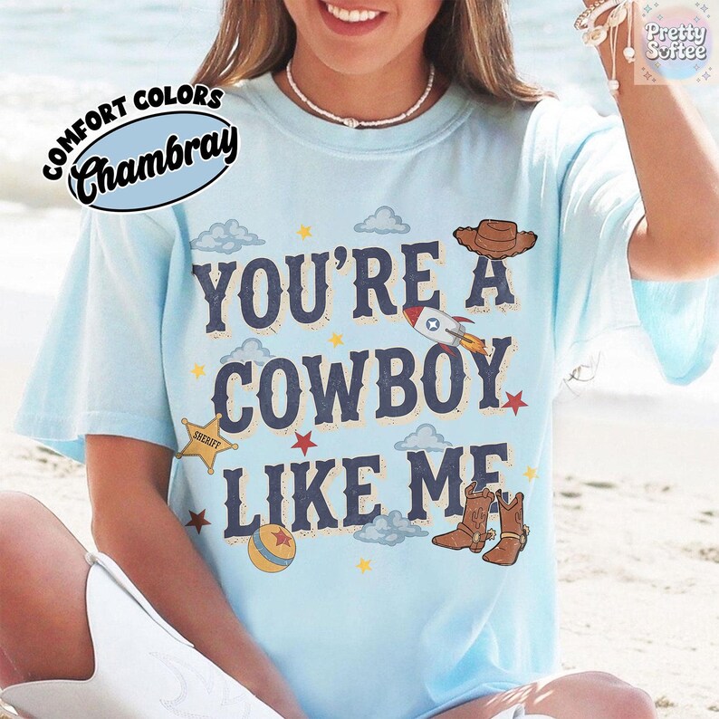 May include: A light blue short-sleeve t-shirt featuring the dark blue text "YOU'RE A COWBOY LIKE ME" surrounded by illustrations of a brown cowboy hat, a rocket ship, a yellow sheriff star, a colorful ball, red stars, and light blue clouds.