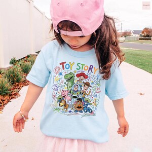 May include: A light blue t-shirt featuring the words "TOY STORY" in colorful letters, with cartoon characters from the movie. The shirt is worn by a child wearing a pink baseball cap.