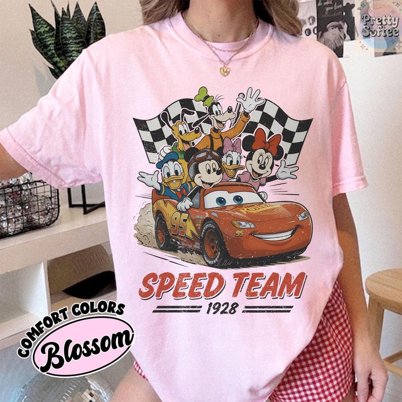 K&ouml;nnte beinhalten: Hellrosa T-Shirt mit einer Grafik von Disney-Figuren in einem Rennwagen. Das Design umfasst Micky Maus, Donald Duck, Goofy, Daisy und Minnie Maus, mit dem Text "SPEED TEAM 1928". Das Shirt ist mit "Comfort Colors Blossom" versehen.