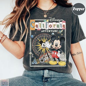 Puede incluir: Camiseta gris oscuro con un gr&aacute;fico colorido de Mickey Mouse saludando frente a una noria, con el texto "Disney California Adventure". La camiseta tiene un aspecto vintage y desgastado.
