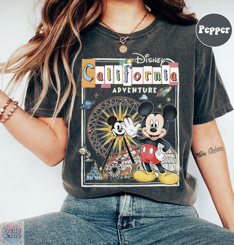 Puede incluir: Camiseta gris oscuro con un gr&aacute;fico colorido de Mickey Mouse saludando frente a una noria, con el texto "Disney California Adventure". La camiseta tiene un aspecto vintage y desgastado.