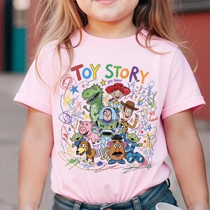 May include: Pink short-sleeved t-shirt featuring a colorful "Toy Story" graphic. The design includes various characters from the animated film, such as Woody, Buzz Lightyear, and Rex, with the words "Toy Story" in a playful font.
