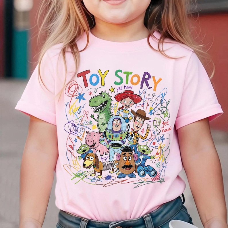 May include: Pink short-sleeved t-shirt featuring a colorful "Toy Story" graphic. The design includes various characters from the animated film, such as Woody, Buzz Lightyear, and Rex, with the words "Toy Story" in a playful font.