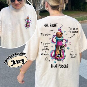 May include: Ivory-colored t-shirt with a graphic of a potion bottle and the text "OH, RIGHT, THAT POISON?" The bottle is pink and blue with a brown cork and contains a black figure. The shirt also has the text "The poison chosen specially to kill kuzco."